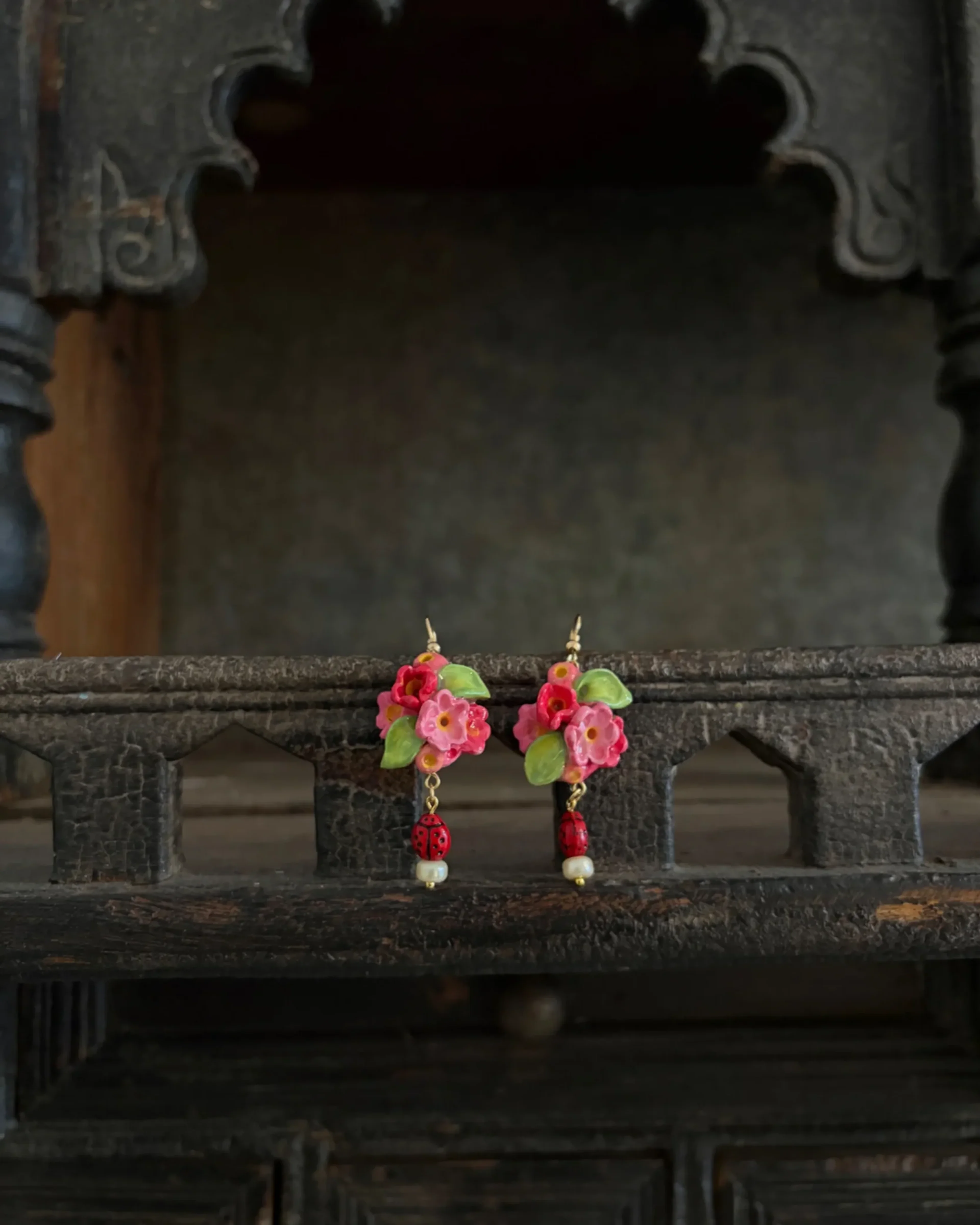 Pink floral cluster earrings with green leaves and ladybird drops, on a carved dark-wood shelf